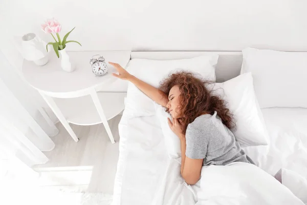 Morning of sleepy young African-American woman turning off alarm clock while lying in bed — Stock Photo, Image