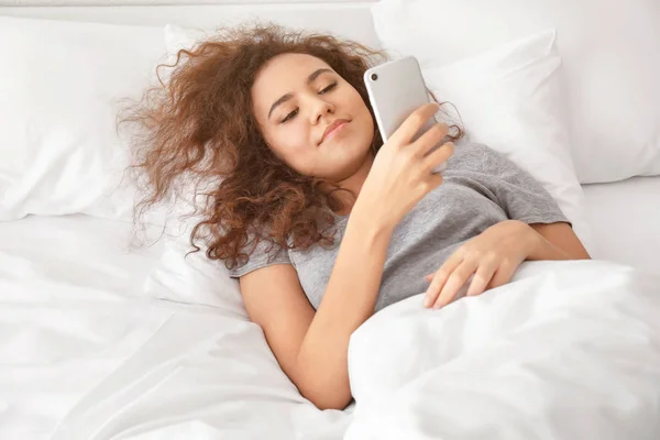 Ráno mladé afroamerické ženy leží v posteli s mobilním telefonem — Stock fotografie