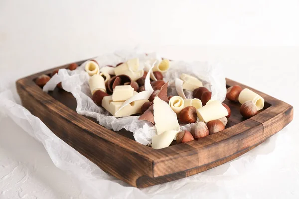 Wooden board with tasty chocolate and hazelnuts on light background — Stock Photo, Image