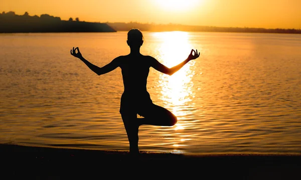 Beautiful young woman practicing yoga near river at sunset — Stock Photo, Image