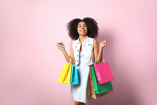 Mooie Afro-Amerikaanse vrouw met boodschappentassen en mobiele telefoon op kleur achtergrond — Stockfoto