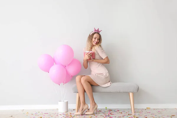 Schöne junge Frau mit Luftballons und Geschenk sitzt in der Nähe von Lichtwand — Stockfoto