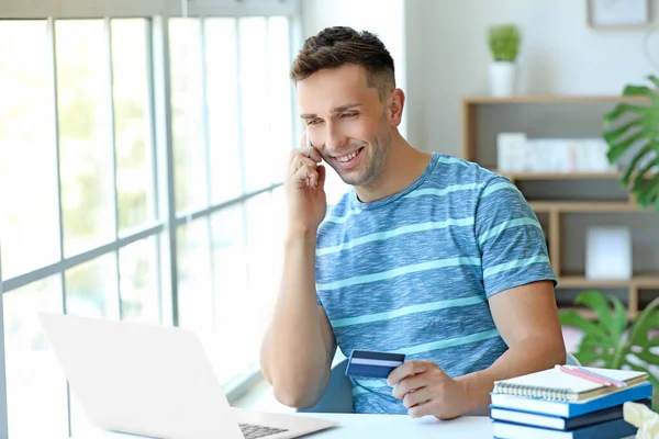 Young man with credit card and laptop talking by mobile phone at home — Stock Photo, Image