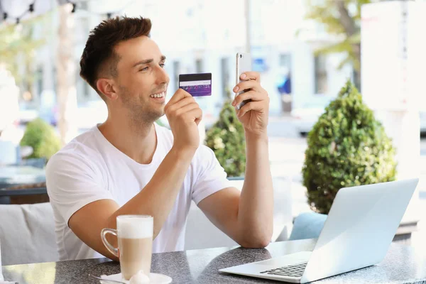 Young man with credit card, mobile phone and laptop sitting in cafe — Stock Photo, Image