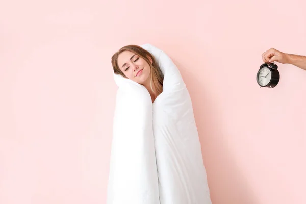 Сонная молодая женщина завернутая в одеяло и мужская рука с будильником на цветном фоне — стоковое фото