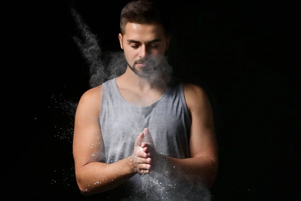 Sporty young man applying talc powder on hands against dark background