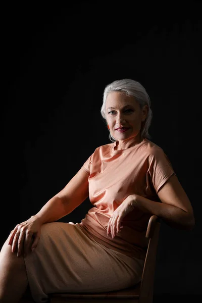 Portrait of stylish mature woman sitting on chair against dark background — Stock Photo, Image