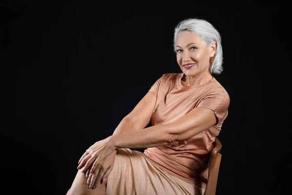 Portrait of stylish mature woman sitting on chair against dark background — Stock Photo, Image