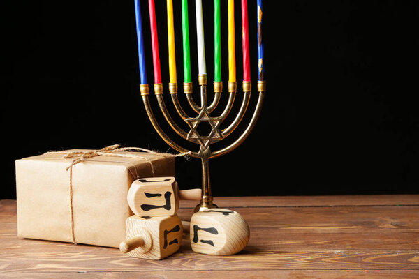 Menorah with candles, dreidels and gift for Hanukkah on table against dark background