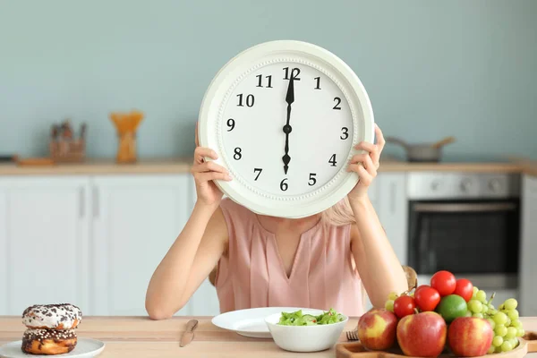 Woman with clock and food sitting in kitchen. Diet concept