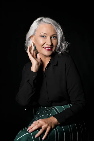 Portrait of stylish mature woman sitting on chair against dark background — Stock Photo, Image