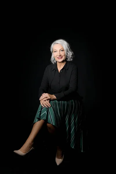 Portrait of stylish mature woman sitting on chair against dark background — Stock Photo, Image