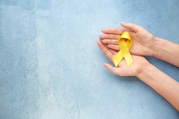 Mãos femininas com fita amarela no fundo da cor. Conceito de consciência do câncer — Fotografia de Stock