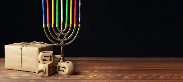 Menorah with candles, dreidels and gift for Hanukkah on table against dark background with space for text — Stock Photo, Image