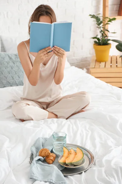 Tray with tasty breakfast and woman reading book on bed — Stock Photo, Image