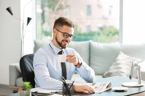 Hombre de negocios guapo bebiendo café mientras lee el periódico en la oficina — Foto de Stock