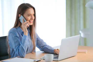 Bilgisayarlı güzel genç masada cep telefonuyla konuşuyor.
