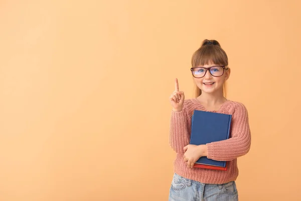 Schattig klein meisje met boeken en opgeheven wijsvinger op kleur achtergrond — Stockfoto