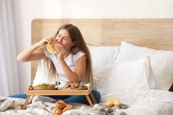 Young woman with addiction to food eating tasty burger on bed at home