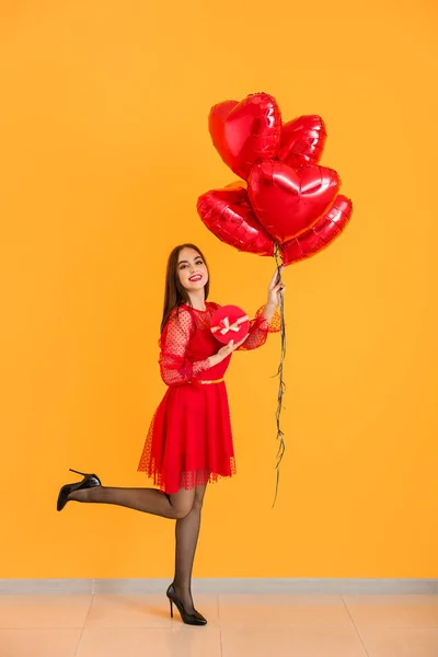 Mulher bonita com balões em forma de coração e presente perto da parede de cor. Celebração do Dia dos Namorados — Fotografia de Stock