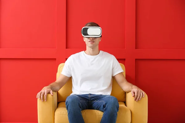 Giovane uomo con occhiali di realtà virtuale seduto in poltrona su sfondo colore — Foto Stock