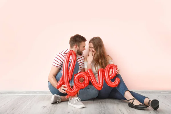 Feliz pareja joven con globo en forma de palabra AMOR sentado cerca de la pared de color. Celebración de San Valentín —  Fotos de Stock