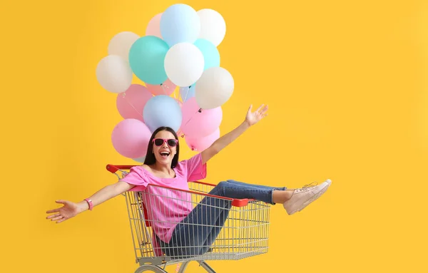 Mujer joven con carrito de compras y globos sobre fondo de color — Foto de Stock