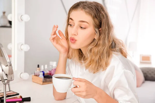 Morning of beautiful young woman drinking coffee while applying makeup in bedroom — Stock Photo, Image