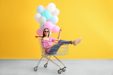 Alışveriş arabası ve renkli duvarın yanında balonları olan genç bir kadın.
