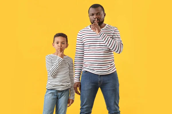 Portrait of African-American man with his little son showing silence gesture on color background — Stock Photo, Image