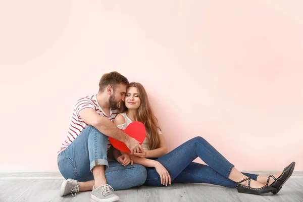 Jovem casal feliz com coração vermelho sentado perto da parede de cor. Celebração do Dia dos Namorados — Fotografia de Stock