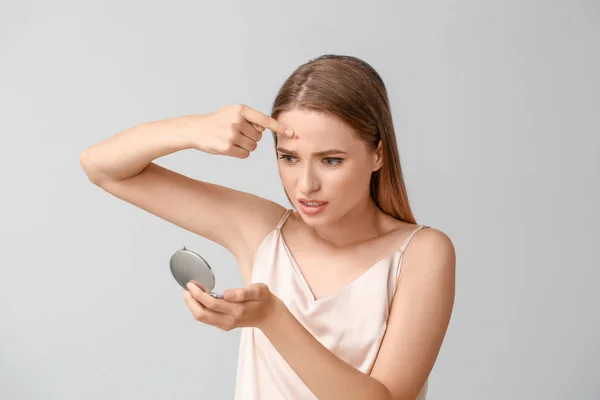 Portrait of young woman with acne problem looking in mirror on light background — Stock Photo, Image