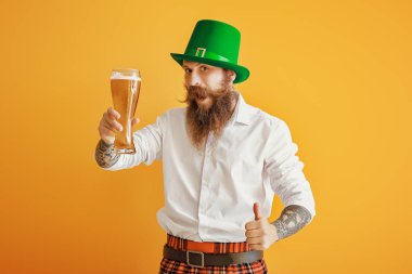 Sakallı adam, elinde bir bardak bira ile arka planda baş parmak hareketi gösteriyor. Aziz Patrick Günü kutlaması
