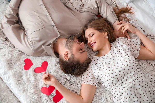 Happy young couple with red hearts lying on bed. Valentine's Day celebration — Stock Photo, Image