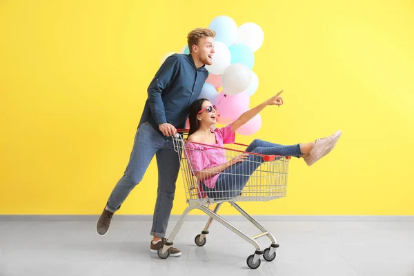 Young couple with shopping cart and balloons near color wall — Stock Photo, Image