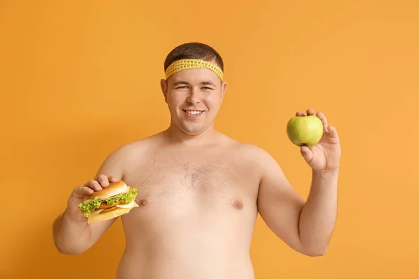 Overweight man with burger and apple on color background. Concept of choice between healthy and unhealthy food