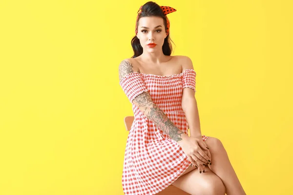 Portrait of beautiful tattooed pin-up woman sitting on chair against color background — Stock Photo, Image