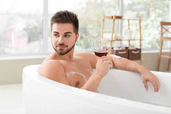 Красивый молодой человек пьет вино, принимая ванну дома — стоковое фото