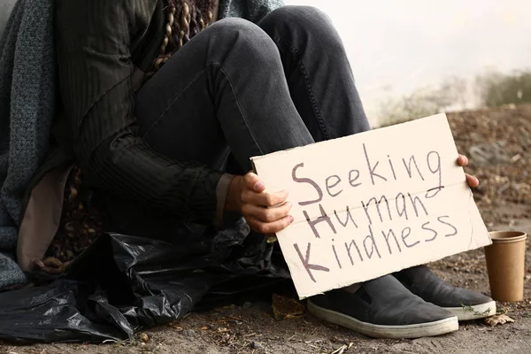 Pobre sem-teto segurando papelão com texto SEEKING HUMAN KINDNESS ao ar livre — Fotografia de Stock