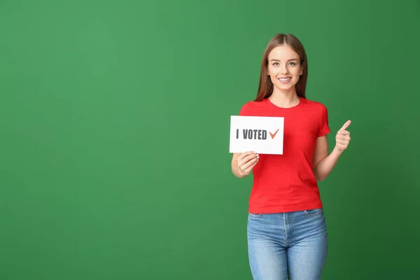 Young woman holding paper with text I VOTED and showing thumb-up on color background — Stock Photo, Image