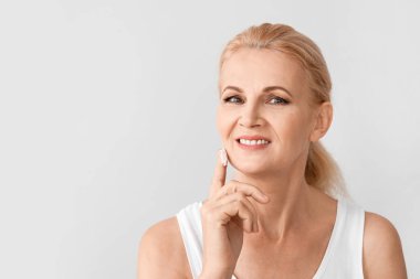Mature woman applying cream on her face against light background