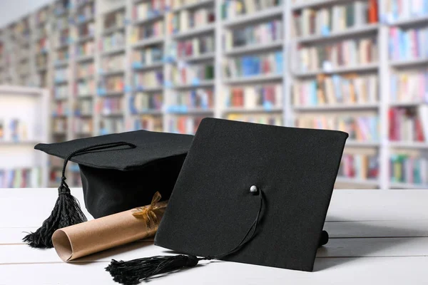 Mortar boards and diploma on table in modern library — Stock Photo, Image