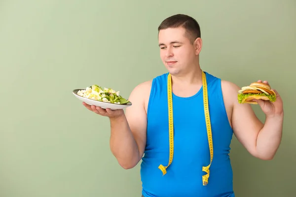 Uomo sovrappeso con metro, insalata e hamburger su sfondo a colori. Concetto di scelta tra alimenti sani e non sani — Foto Stock