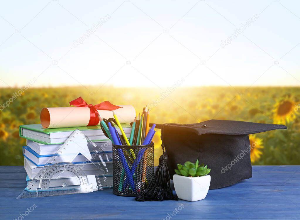 Mortar board, diploma, stationery and books on table near sunflower field
