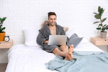 Yatakta dizüstü bilgisayar kullanan bornozlu yakışıklı adam.