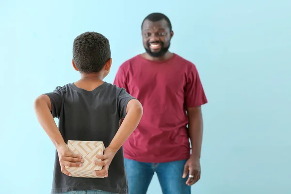 Little African-American boy hiding gift from his father behind back against color background — Stock Photo, Image