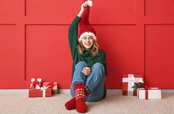 Happy young woman in winter clothes and with Christmas gifts sitting near color wall — Stock Photo, Image
