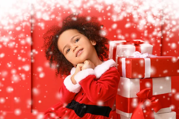 Kis afro-amerikai lány Mikulás jelmez és ajándékok színes háttér hó — Stock Fotó