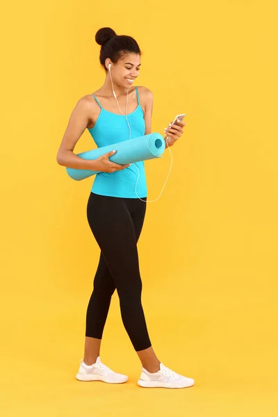 Sporty African-American woman with yoga mat and mobile phone on color background — Stock Photo, Image