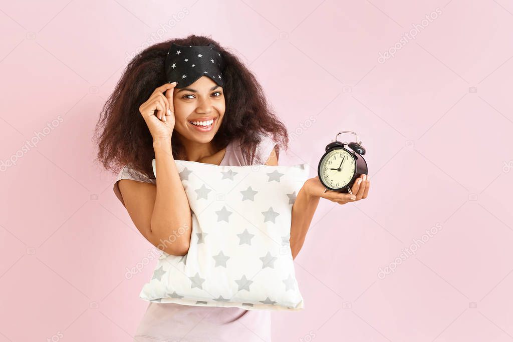Beautiful African-American woman with sleep mask, pillow and alarm clock on color background